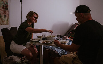 Foto: Markus und Lena Riepl sitzen an einem Tisch und essen mit ihrer Tochter, die im Bett aufrecht sitzt.