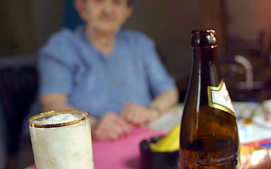 Foto: Am Tisch sitzt eine ältere Frau. Vor ihr steht eine Flasche und ein Glas Bier.