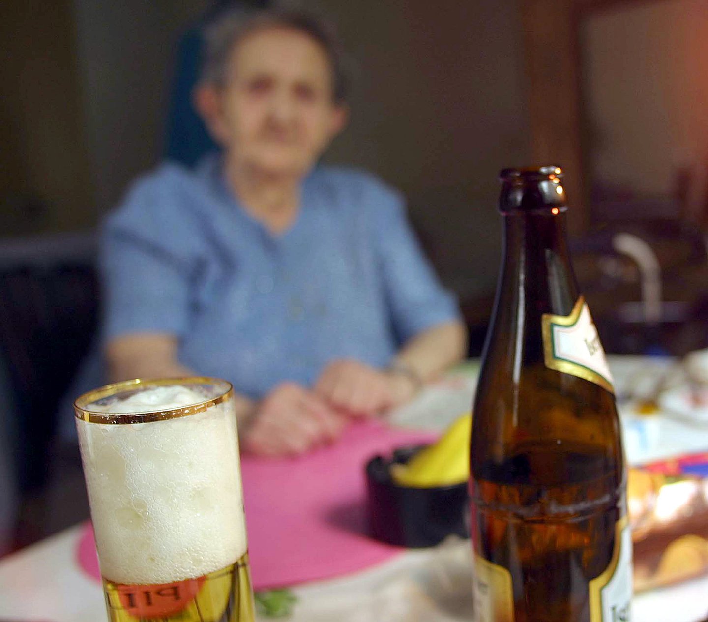 Foto: Am Tisch sitzt eine ältere Frau. Vor ihr steht eine Flasche und ein Glas Bier.