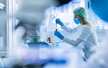 Foto: Eine Wissenschaftlerin mit Schutzmaske, Schutzbrille, Haube und Handschuhen arbeitet mit einer Pipette in einem Labor.