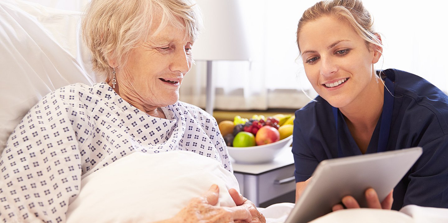 Eine medizinische Kraft zeigt einer älteren Patientin in einem Krankenhausbett Daten auf einem Tablet.