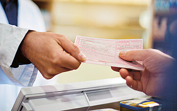 Foto: Ein Arzt oder Apotheker im wei&szlig;en Kittel &uuml;bergibt einer Person eine Verordnung oder ein Rezept.