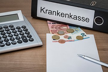Haushalt 2025 Auf einem Schreibtisch liegt ein Ordner mit der Aufschrift 'Krankenkasse', ein Taschenrechner und Geld.