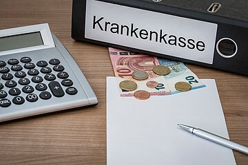Auf einem Schreibtisch liegt ein Ordner mit der Aufschrift 'Krankenkasse', ein Taschenrechner und Geld.