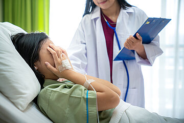 Eine Frau liegt im Krankenhaus im Bett. Sie h&auml;lt sich beide H&auml;nde vors Gesicht, an ihrer rechten Hand ist ein Zugang mit Tropf sichtbar. Auf der anderen Seite des Bettes steht eine &Auml;rztin, die sich ihr zugewandt hat.