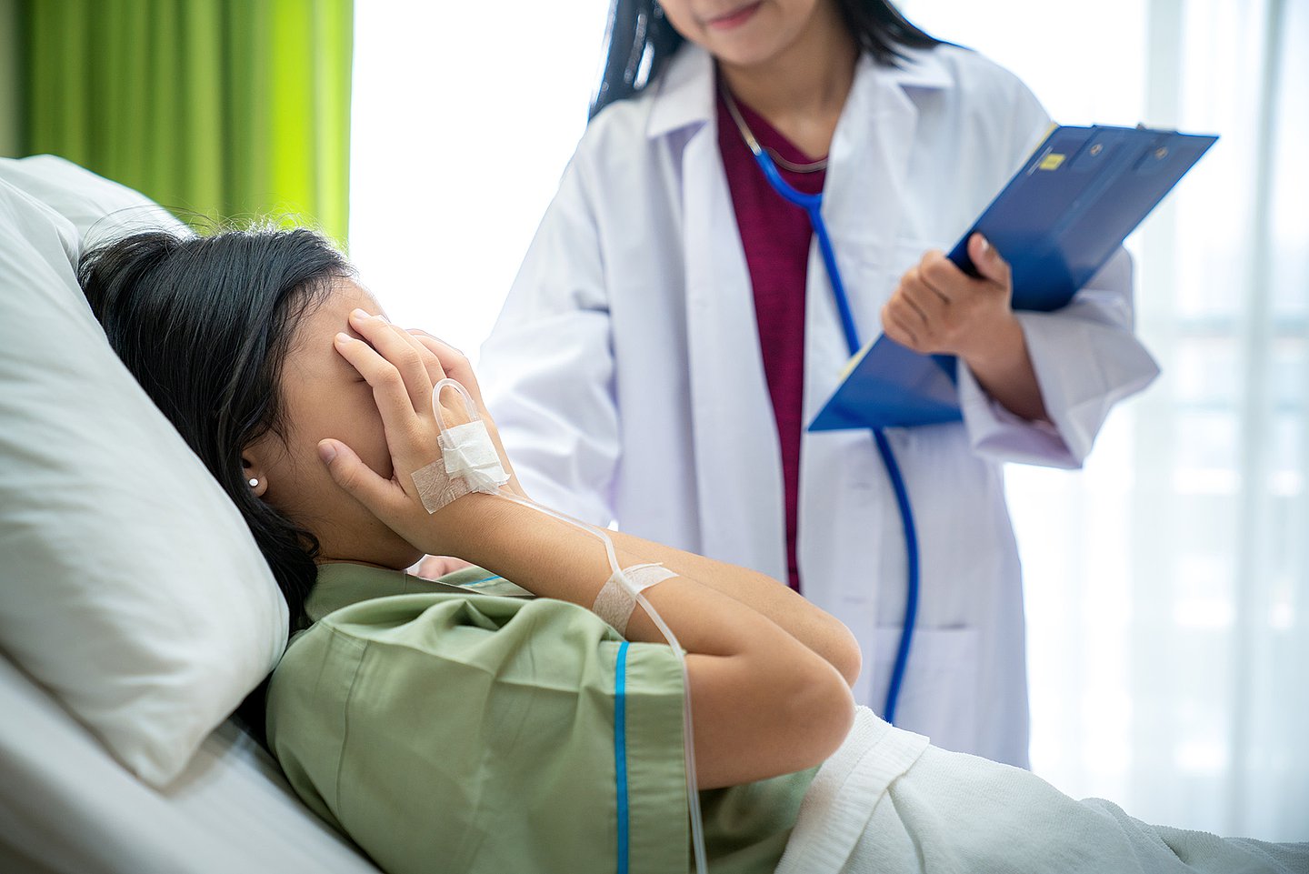 Eine Frau liegt im Krankenhaus im Bett. Sie hält sich beide Hände vors Gesicht, an ihrer rechten Hand ist ein Zugang mit Tropf sichtbar. Auf der anderen Seite des Bettes steht eine Ärztin, die sich ihr zugewandt hat.