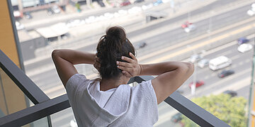 Foto: Eine Frau steht auf einem Balkon, unter ihr fahren Autos. Sie h&auml;lt sich die Ohren zu.