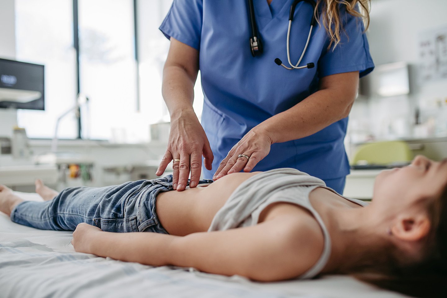 Das Foto zeigt ein Kind, das auf einer Untersuchungsliege im Krankenhaus liegt und von einer Ärztin im Bauchbereich untersucht und abgetastet wird.