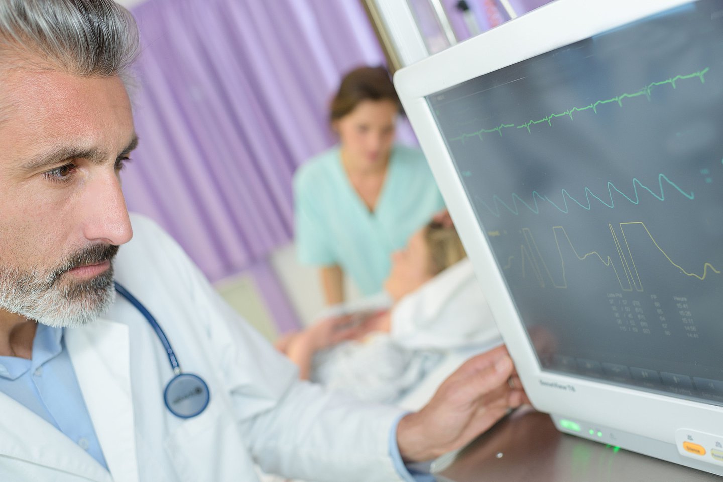 Ein Arzt schaut auf einen Monitor mit Vitaldaten eines Patienten, im Hintergrund kümmert sich eine Krankenschwester um eine im Krankenbett liegende Person.