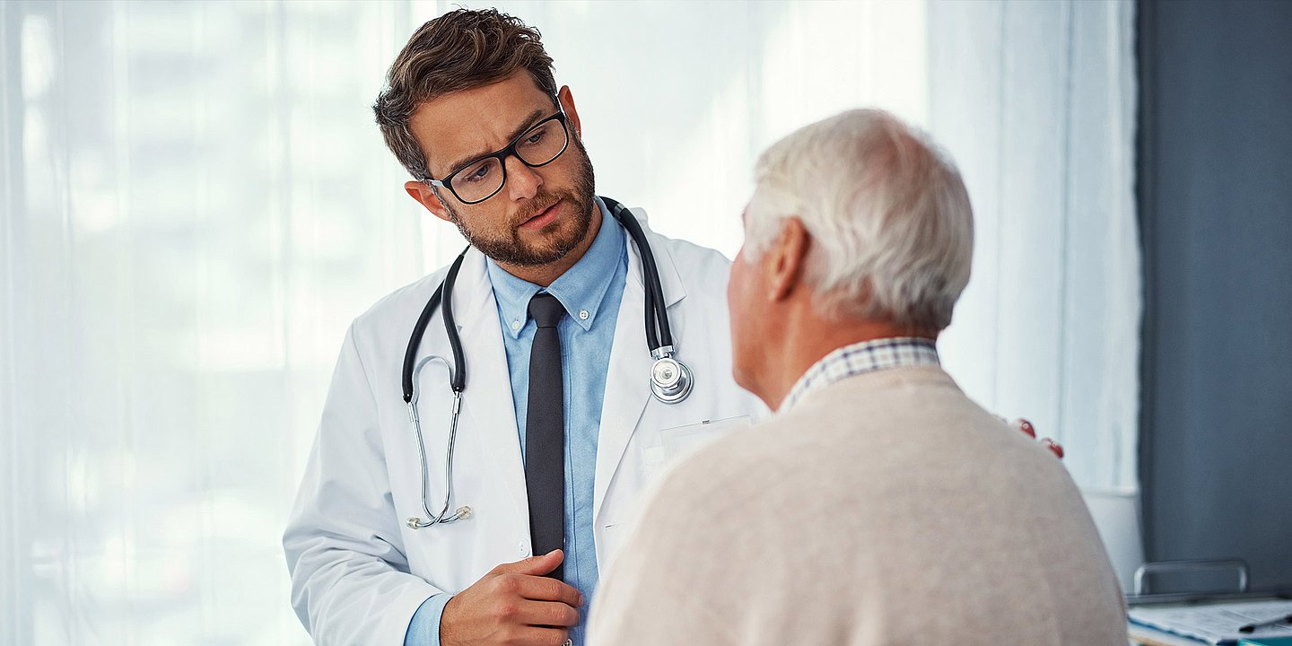Ein junger Arzt im weißen Kittel spricht mit einem älteren Herrn, der von hinten zu sehen ist.