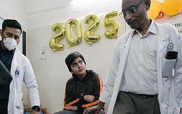 Im Wartezimmer sitzt ein Junge auf einer Liege und zwei Ärzte stehen neben ihm. Einer von ihnen ist Dr. Mathew Varghese, der ihm Übungen gegen die Klumpfüße zeigt.