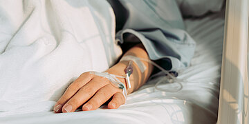 Foto: Hand einer Patientin oder eines Patienten im Krankenhausbett mit liegendem Infusionszugang.