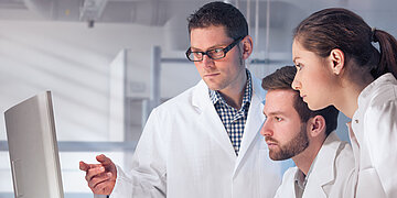 Ein Besprechungsraumim Krankenhaus. Ein Team aus &Auml;rzten schaut gemeinsam auf den Bildschirm. Dabei zeigt ein Arzt auf etwas, was auf dem Monitor dargestellt wird.