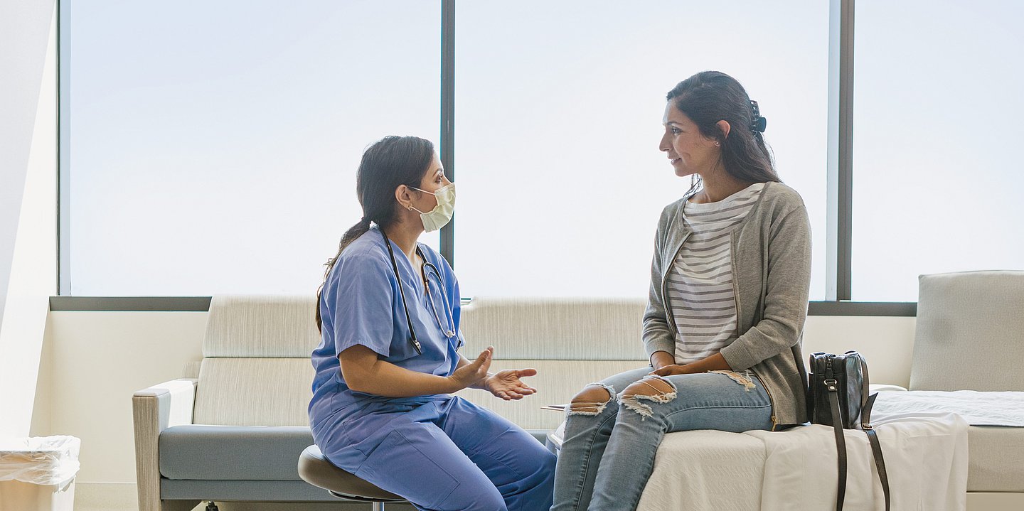 Auf einem Krankenhausbett sitzt eine Patientin. Vor ihr sitzt eine Krankenpflegerin mit Mundschutz in Arbeitskleidung und erklärt der Patientin etwas.