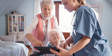 Foto: Eine Pflegekraft zeigt zwei älteren Damen, eine davon deutlich jünger, eine älter und im Rollstuhl, etwas auf einem Tablet.