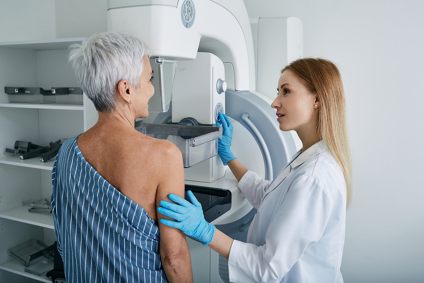 Foto: ältere Frau von hinten, die gerade ein Mammographie-Screening macht. Neben ihr eine radiologische Fachkraft, die das Gerät bedient.