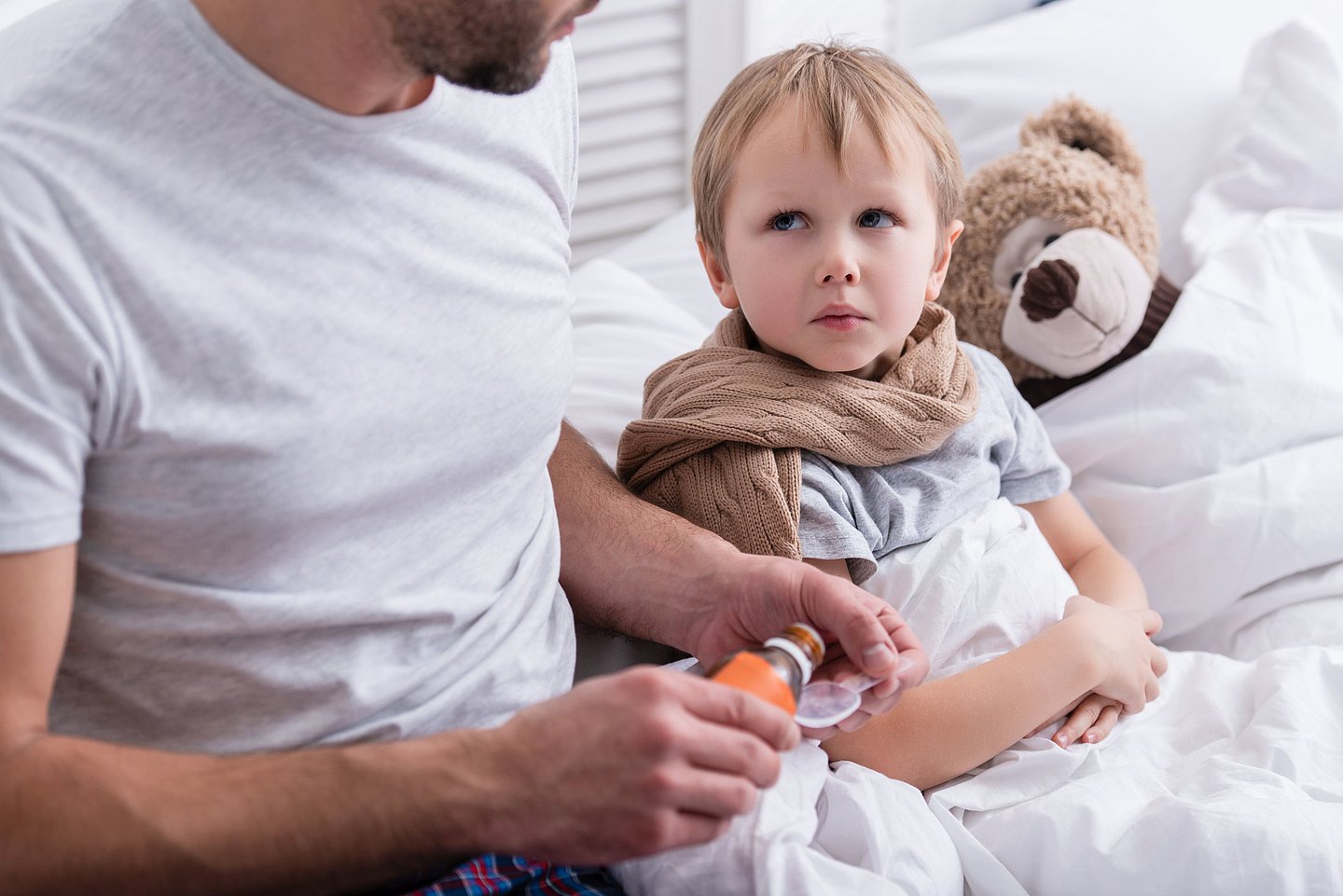 Foto zeigt einen Vater, der am Bett seines kranken Kindes sitzt und diesem einen medizinischen Saft verabreicht. 