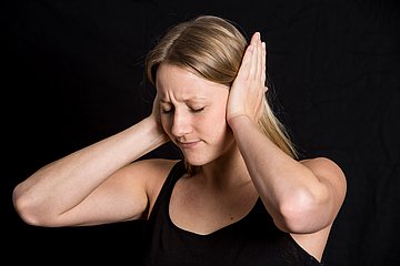 Das Foto zeigt eine Frau, die sich mit beiden Händen und schmerzverzerrtem Gesicht die Ohren zuhält.