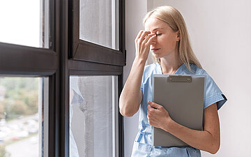 Eine ersch&ouml;pfte junge Pflegeperson steht am Fenster und h&auml;lt einen Augenblick inne.