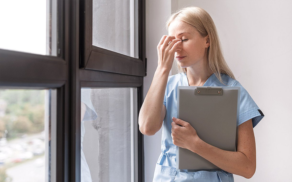 Eine erschöpfte junge Pflegeperson steht am Fenster und hält einen Augenblick inne.