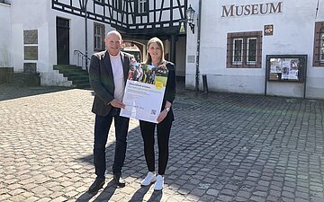 J&uuml;rgen Dreher und Katharina Gutjahr von der AOK S&uuml;dlicher Oberrhein stehen in einer historischen Umgebung und pr&auml;sentieren ein Plakat.