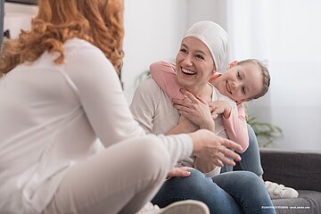 Krebspatientin mit M&auml;dchen, das sie umarmt und einer weiteren Frau auf der Couch