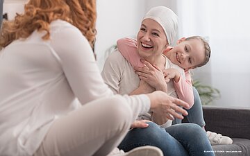 Eine an Krebs erkrankte Frau mit einem Mädchen, das sie umarmt und einer weiteren Frau auf der Couch