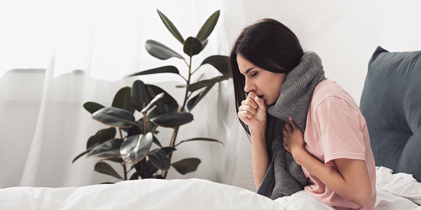 Frau sitzt im Bett mit Schal und hustet in ihre Faust.