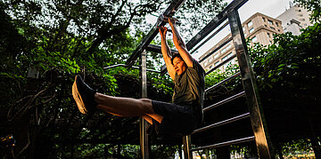 Ein Mann macht eine Beinhebe&uuml;bung h&auml;ngend an einer Klimmzugstange in einem gr&uuml;nen Stadtpark.