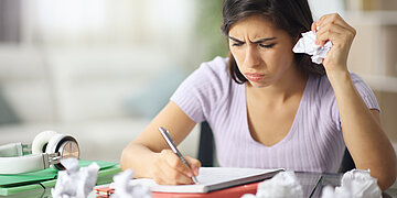 Foto einer jungen Frau, die angestrengt schreibt. Um sie herum liegen viele zerkn&uuml;llte Bl&auml;tter.