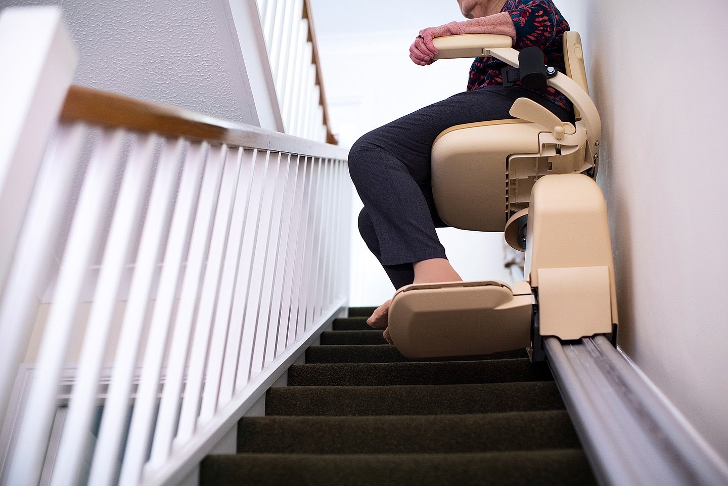 Vom unteren Ende einer Treppe sieht man nach oben auf einen Treppenlift in dem eine ältere weibliche Person sitzt und zum oberen Stockwerk fährt.