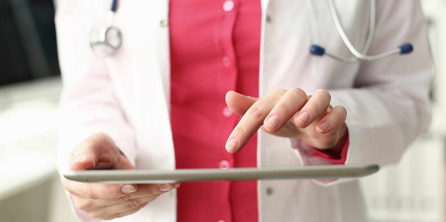 Foto: Ein Mensch im weißen Arztkittel hält ein Tablet in der Hand und tippt mit einem Finger.