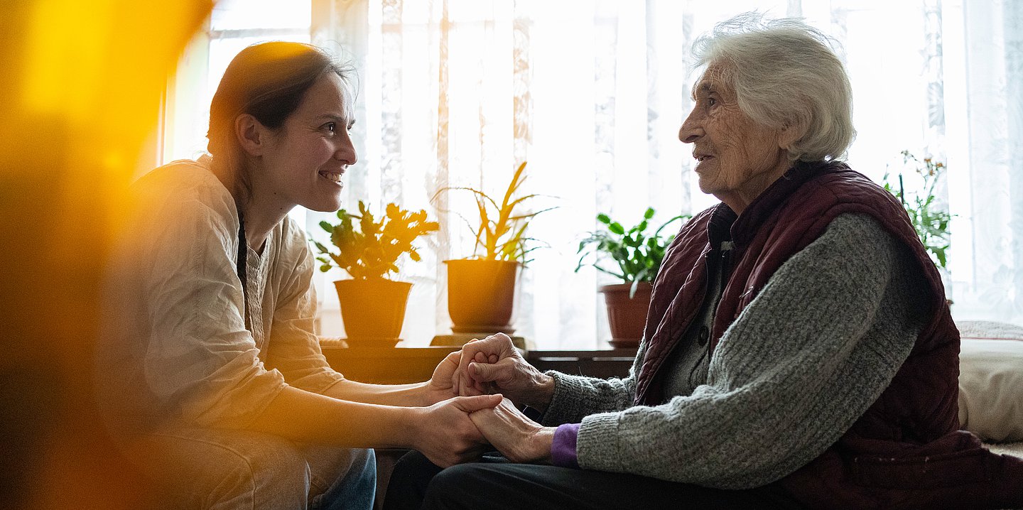 Eine Pflegekraft sitz einer älteren Dame gegenüber und hält während einer Unterhaltung ihre Hände.
