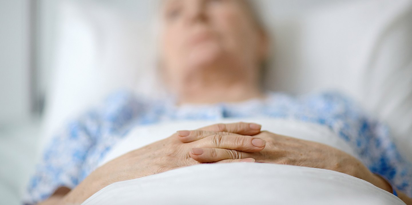 Das Foto zeigt eine alte SEniorin im Bett (Weichzeichner). Die Hände liegen gefaltet auf der weißen Bettdecke.