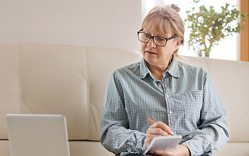Selbsthilfe im Wandel Foto einer älteren Dame, die vom heimischen Sofa aus interessiert auf ihren Laptop-Bildschirm blickt.