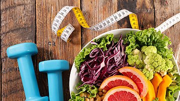 Auf einem Holztisch steht eine Schale mit frischen Salat. Daneben liegen ein Maßband und Hanteln.