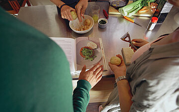 Einige Personen stehen um ein Kochbuch und bereiten gemeinsam Speisen zu. Sie sind aus der Vogelperspektive zu sehen.