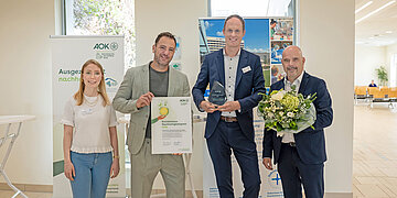 Vier Personen stehen nebeneinander und blicken in die Kamera. Links ist weiblich, die anderen drei sind m&auml;nnlich. Der Mann ganz rechts h&auml;lt einen gro&szlig;en Blumenstrau&szlig; in der Hand. Der rechts daneben einen Preis und der zweite von links ein Zertifikat mit der Aufrschrift "Krankenhaus Nachhaltigkeitspreis 2026". 