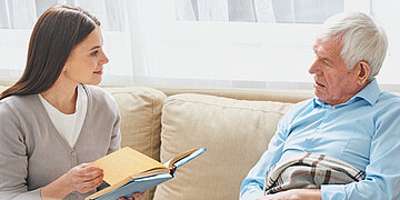 Foto: Eine junge Frau sitzt mit einem &auml;lteren Mann auf dem Sofa und h&auml;lt ein Buch in der Hand. Sie schauen sich aufmerksam an.