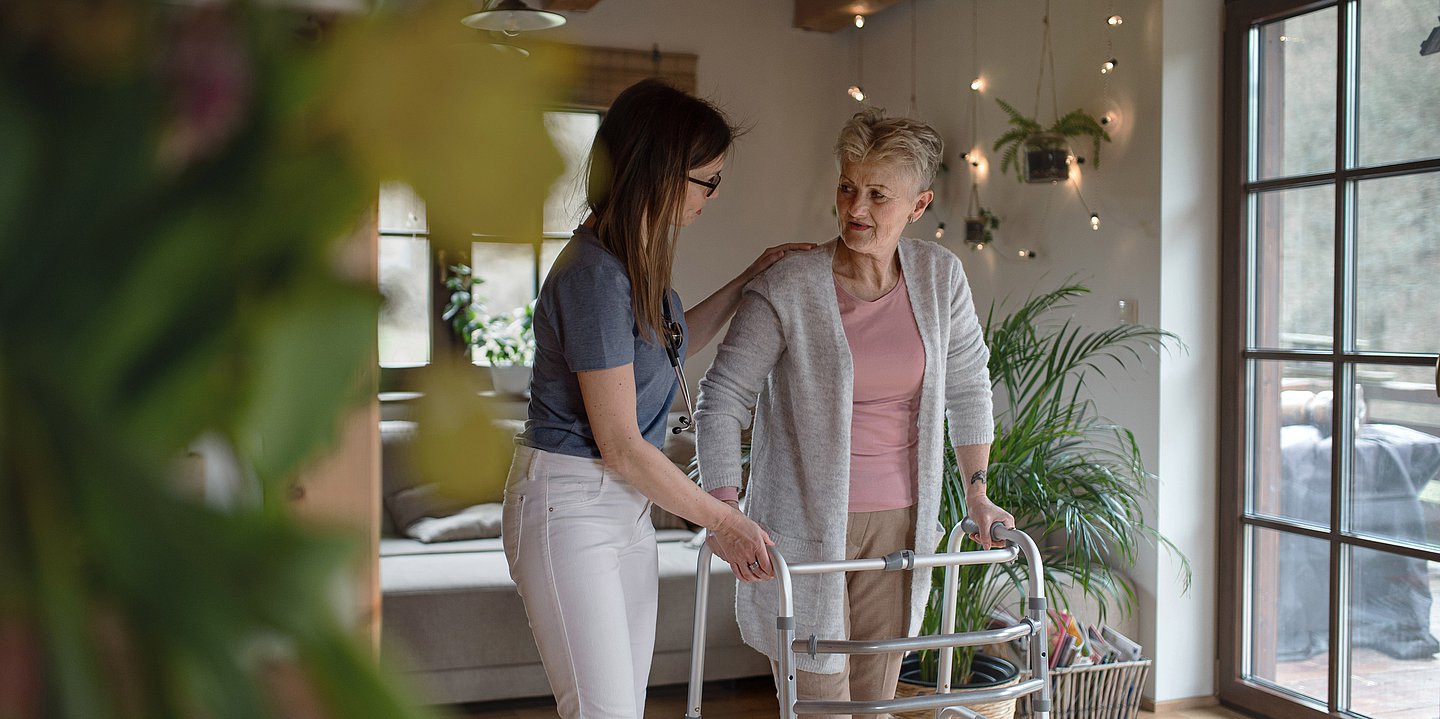Foto einer jungen Frau, die in einem Wohnzimmer einer alten Dame am Rollator hilft