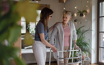 Doppelbelastung von Frauen Foto einer jungen Frau, die in einem Wohnzimmer einer alten Dame am Rollator hilft
