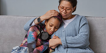 Eine Mutter sitzt auf der Couch und h&auml;lt ihre neben ihr sitzende Teenager-Tochter im Arm. 