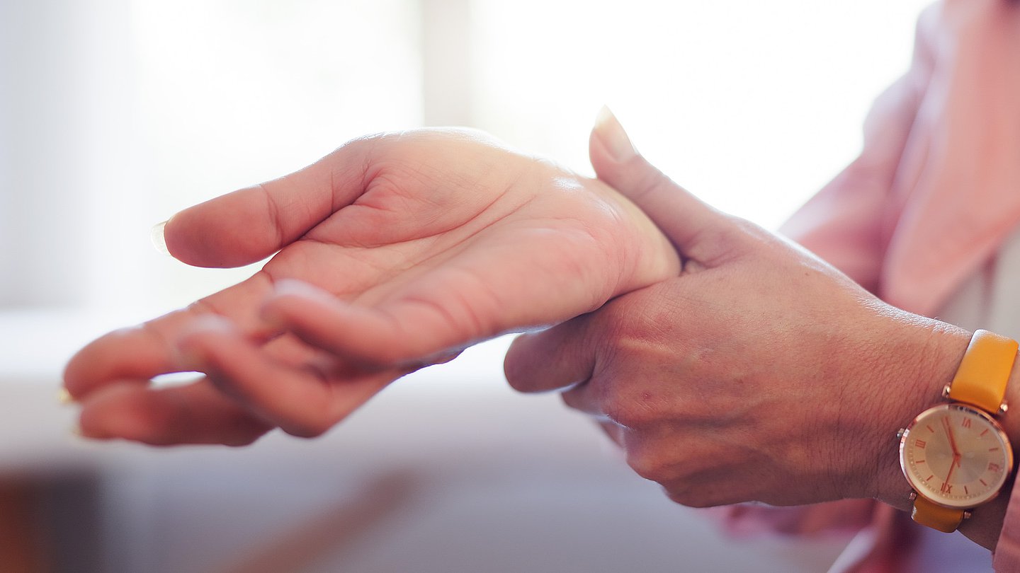 Eine ältere Frau in Nahaufnahme fasst mit der linken Hand an ihr rechtes Handgelenk.