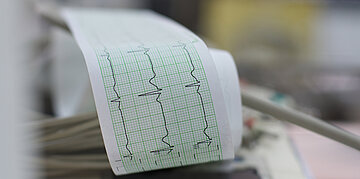 Ein EKG ist in Betrieb. Auf dem Ausdruck sind auf gr&uuml;nem Milimeterpapier schwarze Zick-Zack-Kurven abgebildet, die den Herzschlag darstellen.