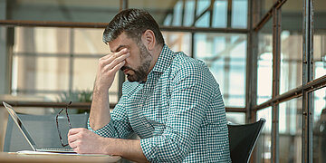 Ein Mann in gr&uuml;n-kariertem Hemd sitzt im B&uuml;ro vor seinem Laptop. Sein Kopf ist gesenkt. Er hat seine Brille in der linken Hand, die Rechte Hand bedeckt seine Augen. 