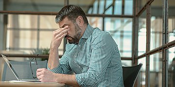 Ein Mann in grün-kariertem Hemd sitzt im Büro vor seinem Laptop. Sein Kopf ist gesenkt. Er hat seine Brille in der linken Hand, die Rechte Hand bedeckt seine Augen.
