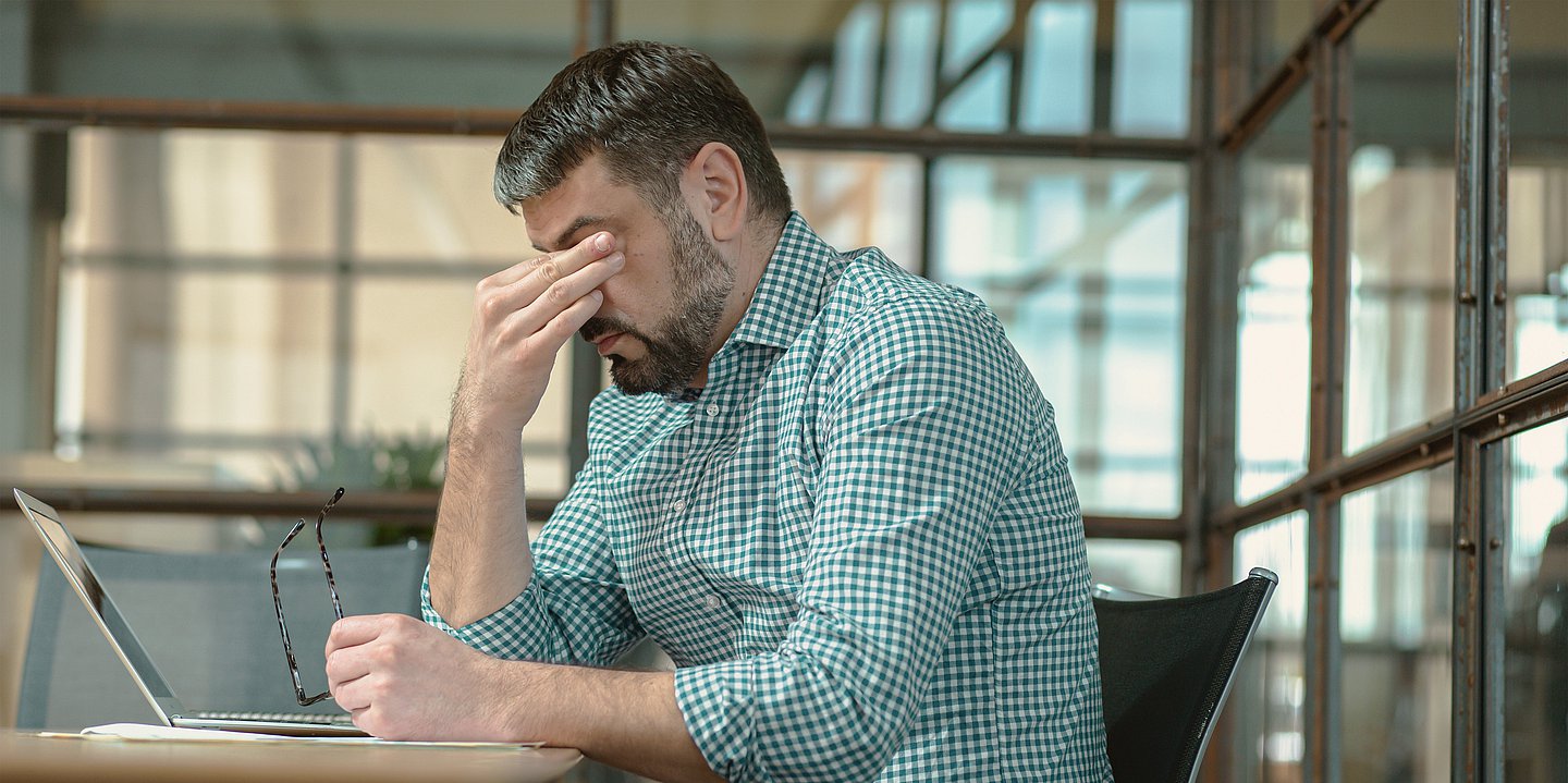 Ein Mann in grün-kariertem Hemd sitzt im Büro vor seinem Laptop. Sein Kopf ist gesenkt. Er hat seine Brille in der linken Hand, die Rechte Hand bedeckt seine Augen. 