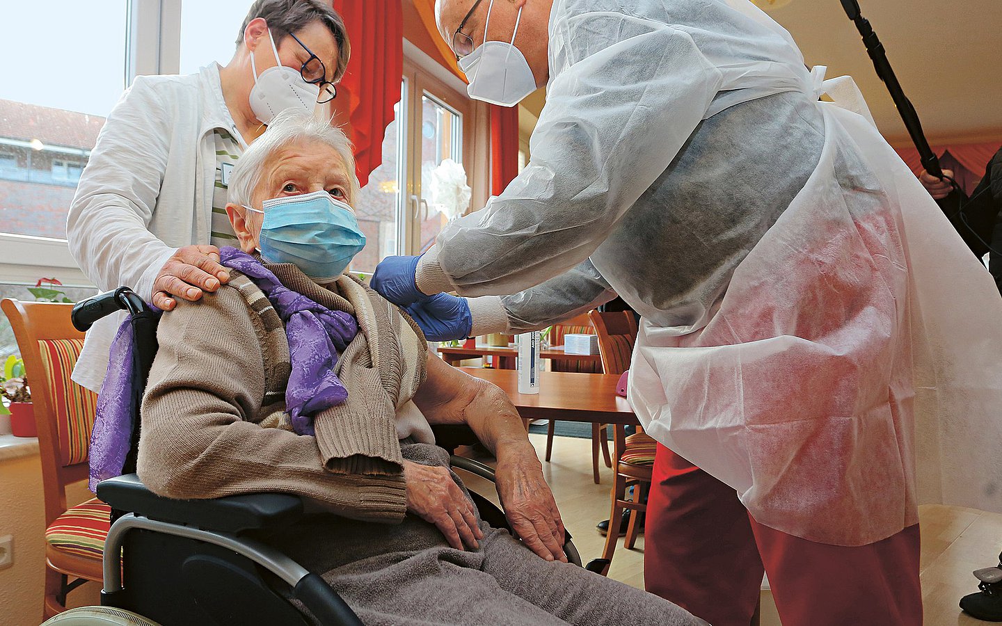 Foto einer alten Frau mit Maske im Pflegeheim. Um sie herum sind Helfende in Schutzkleidung, die sie impfen