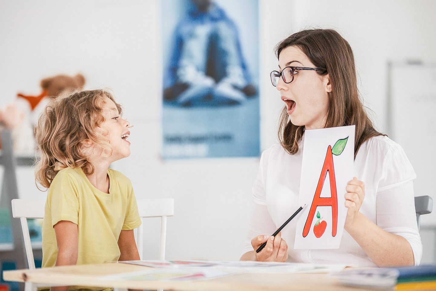 Sprachtherapeutin hält ein Blatt mit dem Buchstaben 'A' hoch und übt mit einem Jungen die Aussprache
