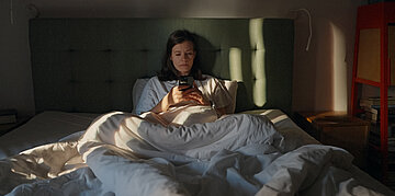 In einem abgedunkelten Zimmer sitzt eine Frau im Bett und schaut in ihr Handy.